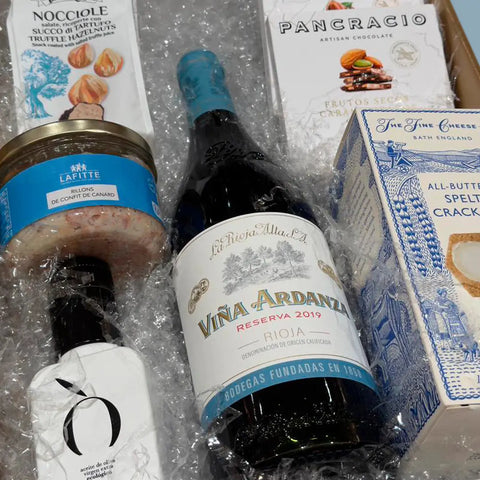 caja de regalo con una botella de vino, una botella de aceite de oliva extra virgen, rillons de confit de canard, avellanas con trufa, crackers con spelta y chocolate Pancracio sobre viruta blanca dentro de una caja dorada.