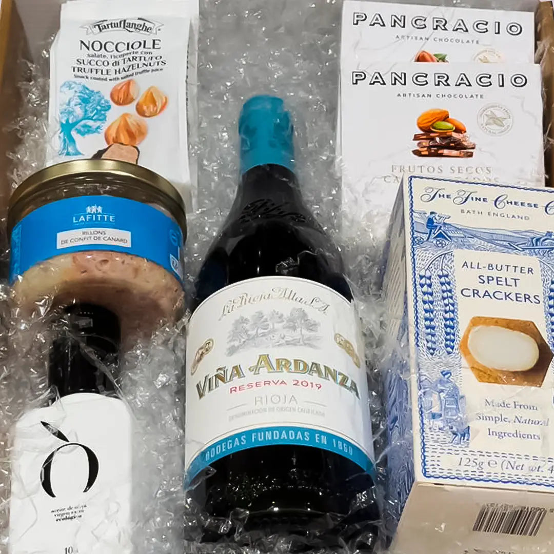 caja de regalo con una botella de vino, una botella de aceite de oliva extra virgen, rillons de confit de canard, avellanas con trufa, crackers con spelta y chocolate Pancracio sobre viruta blanca dentro de una caja dorada.
