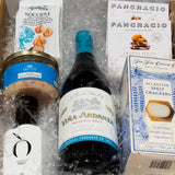 caja de regalo con una botella de vino, una botella de aceite de oliva extra virgen, rillons de confit de canard, avellanas con trufa, crackers con spelta y chocolate Pancracio sobre viruta blanca dentro de una caja dorada.